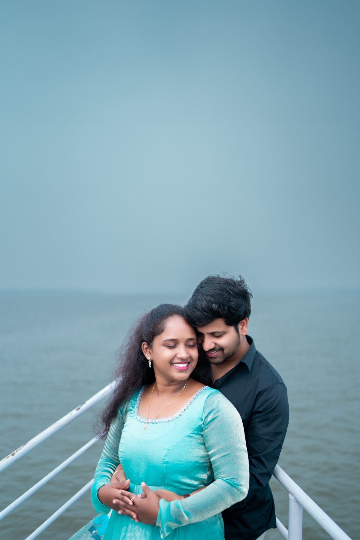 Couple embracing on a jetty over calm grey water during an overcast pre-wedding shoot