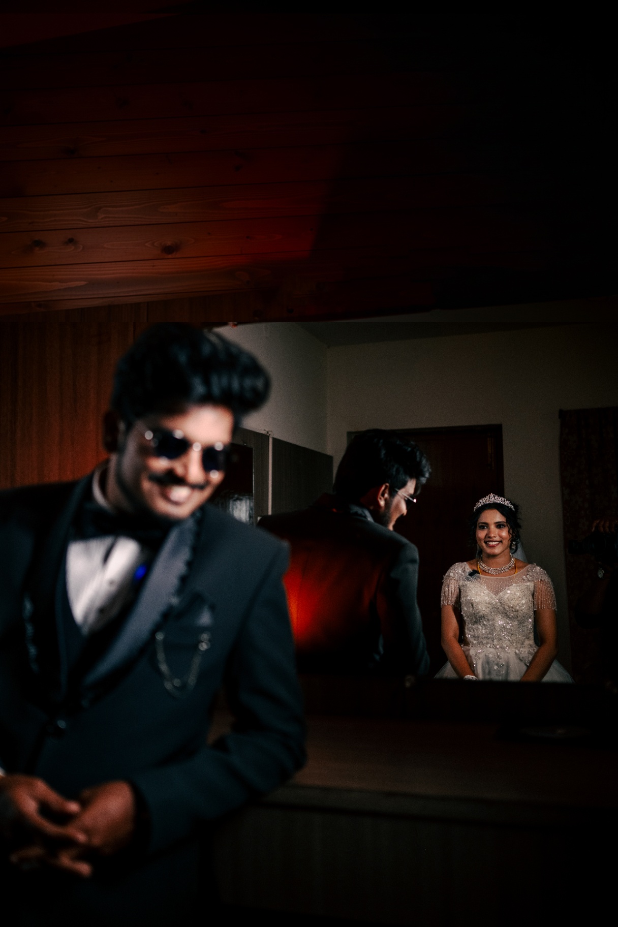 Groom in the foreground with the bride reflected in a mirror behind him
