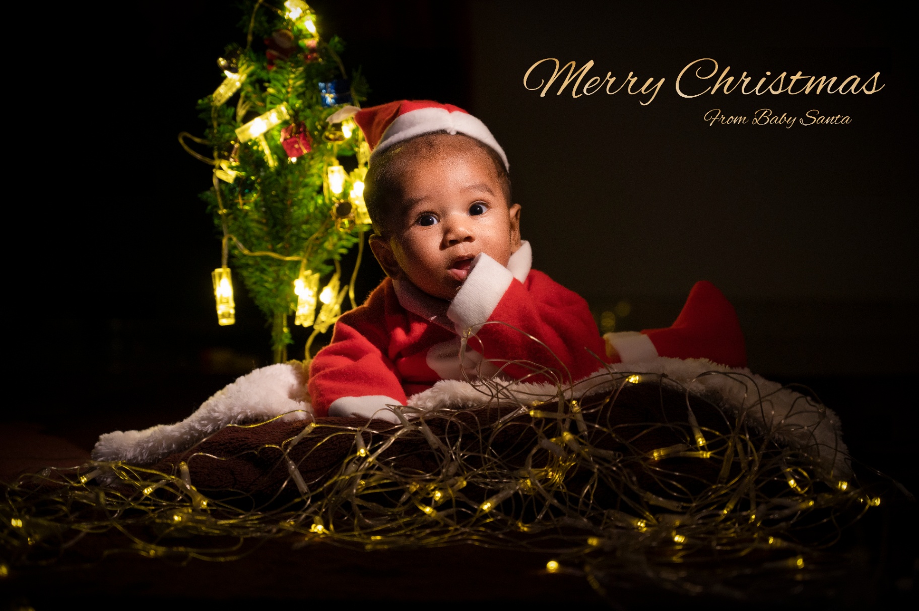 Baby in a Santa outfit with festive lights and a Christmas tree