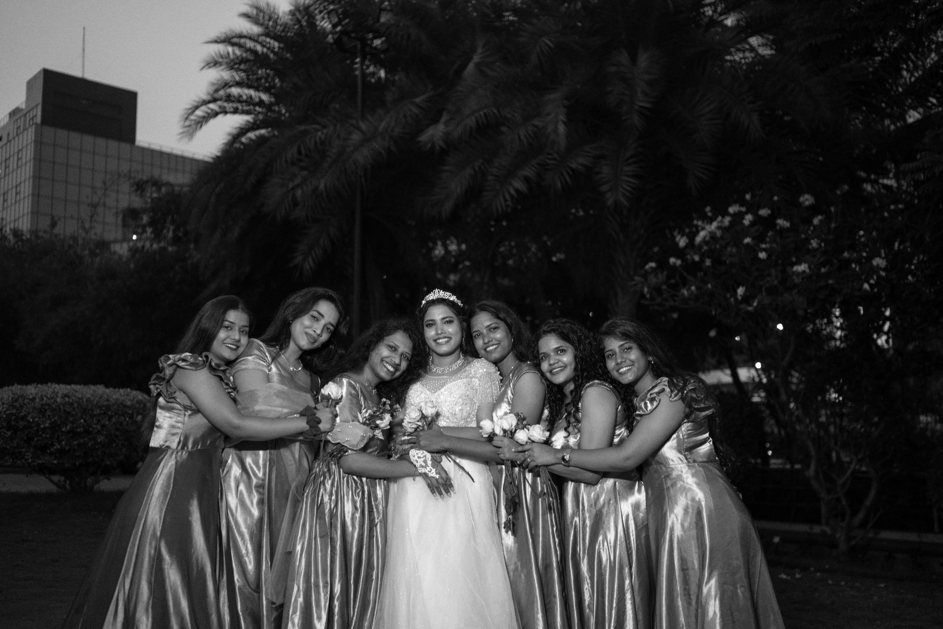 Black and white group portrait of a bride surrounded by bridesmaids outdoors at dusk